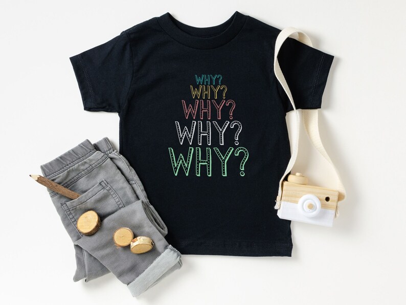 Op de afbeelding: Een zwart T-shirt met het woord "WHY?" vijf keer herhaald in een regenboog van kleuren. Het shirt ligt plat op een wit oppervlak met een grijze spijkerbroek en een houten speelgoedcamera.