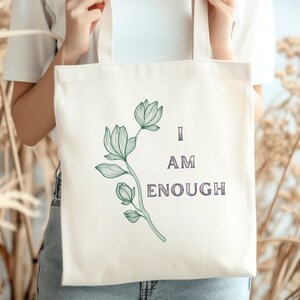 May include: Cream-colored tote bag featuring a green floral design and the words "I AM ENOUGH" in a dark purple font. The bag is held up against a backdrop of dried plants.
