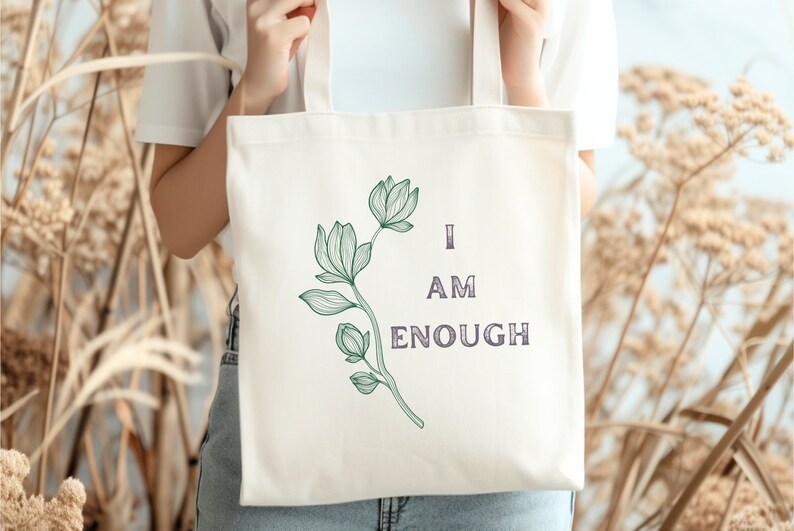 May include: Cream-colored tote bag featuring a green floral design and the words "I AM ENOUGH" in a dark purple font. The bag is held up against a backdrop of dried plants.