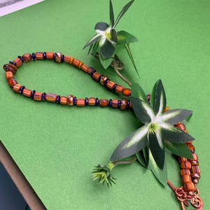 May include: A necklace made of orange and blue glass beads with a copper clasp. The necklace is lying on a green surface with two small green plants.
