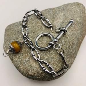 May include: A silver chain bracelet with a toggle clasp and a single amber-colored bead. The bracelet is on a gray rock.