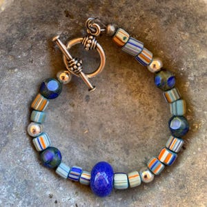 May include: A bracelet featuring a variety of glass beads in shades of blue, orange, and white, interspersed with metallic silver-toned beads. The piece is secured with a silver-toned toggle clasp, showcasing a mix of striped and solid bead designs.