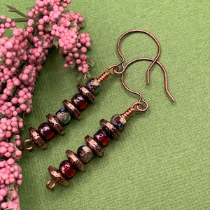 May include: A pair of handcrafted earrings with copper-colored ear wires. Each earring features a series of dark red and multicolored beads, separated by textured, copper-toned metal discs. The earrings are displayed on a green surface, with pink floral accents.