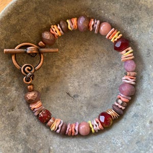 May include: A beaded bracelet featuring a copper toggle clasp with a spiral design. The bracelet is composed of various beads in shades of pink, red, orange, and brown, including faceted glass beads, smooth round stones, and thin, wavy disc-shaped spacers.