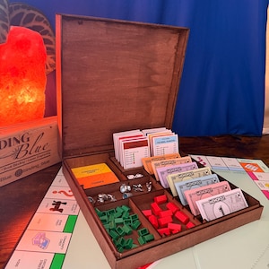 May include: A dark brown wooden board game organiser box with an open lid, containing organised compartments for paper money, property cards, green houses, red hotels, and metal game tokens, resting on a Monopoly game board surface.