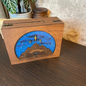 May include: A wooden box with a hinged lid. The lid is engraved with the words "You're my sunshine". The box has a blue and white image of a mountain range with stars in the sky.