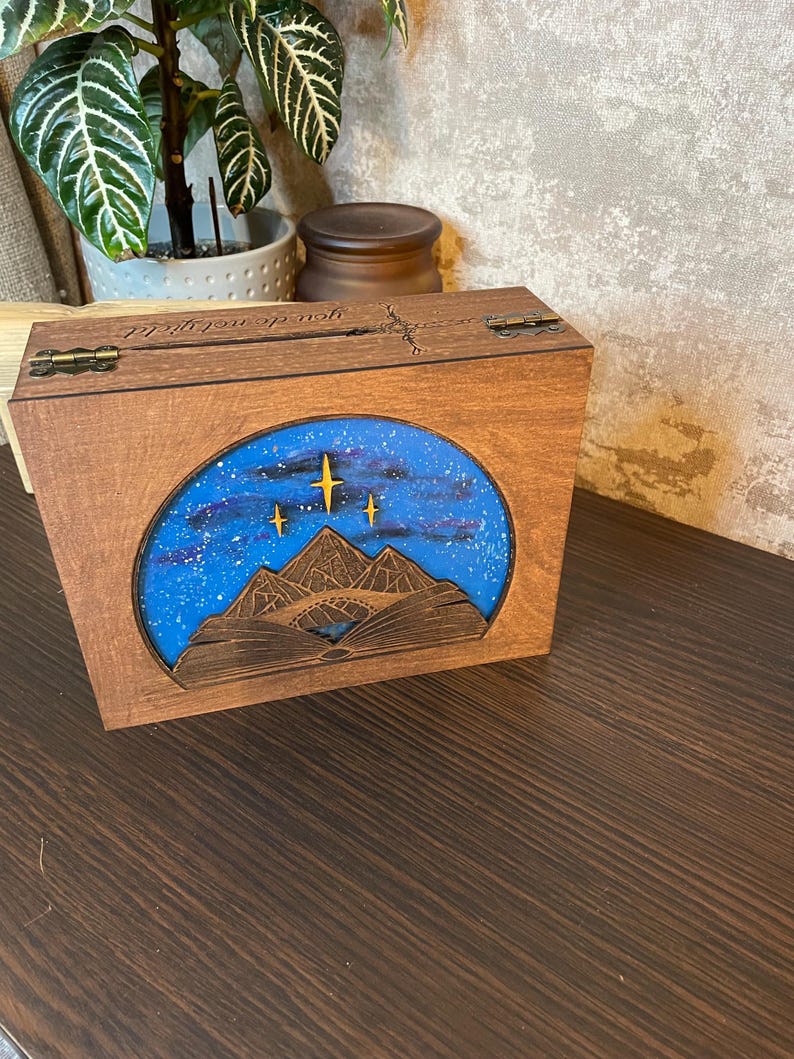 May include: A wooden box with a hinged lid. The lid is engraved with the words "You're my sunshine". The box has a blue and white image of a mountain range with stars in the sky.
