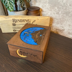 May include: A wooden box with a blue and white starry sky scene inside. The box has a crescent moon with the words "light it up" carved into the wood.