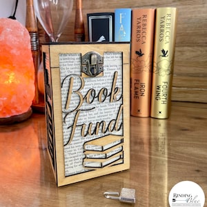 May include: A wooden box with a lock and key, decorated with a book-shaped cutout and the words "Book Fund". The box is sitting on a wooden surface with a salt lamp and a stack of books in the background.