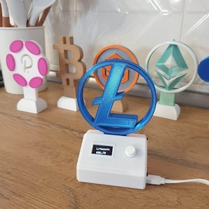 Może przedstawiać: Biały wyświetlacz kryptowaluty Litecoin wydrukowany w 3D z niebieskim logo Litecoin i małym wyświetlaczem cyfrowym pokazującym cenę Litecoin, 92,79 €.