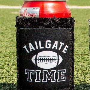 Może przedstawiać: Czarno-biały ochładzacz do puszek z napisem "TAILGATE TIME" i białą grafiką piłki nożnej. Chłodziarka wykonana jest z miękkiej, czarnej tkaniny z błyszczącym wykończeniem.
