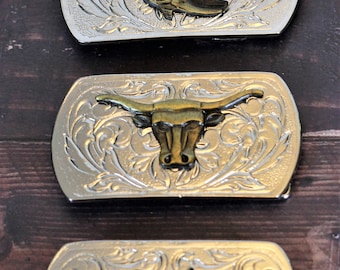 May include: Three silver belt buckles with different western-themed designs. The first buckle features a pair of cowboy boots, the second buckle features a bull's head, and the third buckle features a saddle.
