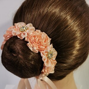 May include: A peach-coloured hair accessory with fabric flowers and sparkling rhinestone accents. The flowers are arranged in a circular pattern, with a ribbon tie. Designed to be worn around a bun.
