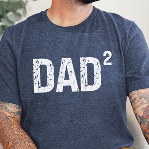 May include: Navy blue t-shirt with the white text "DAD²" in a distressed font. The shirt is a crew neck and appears to be made of a soft material. The shirt is a casual style.