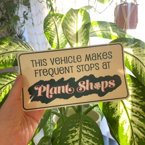 May include: A beige sign with the text "This vehicle makes frequent stops at Plant Shops" in a retro font. The word "Plant" is in green and the word "Shops" is in green with a pink leaf icon.