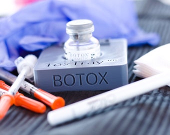 May include: A gray plastic tray with the word "BOTOX" printed on it. A clear glass vial with a blue label that says "BOTOX COSMETIC" is sitting in the tray. There are also several orange and clear syringes, a white cotton swab, and a stack of white paper squares on the tray.