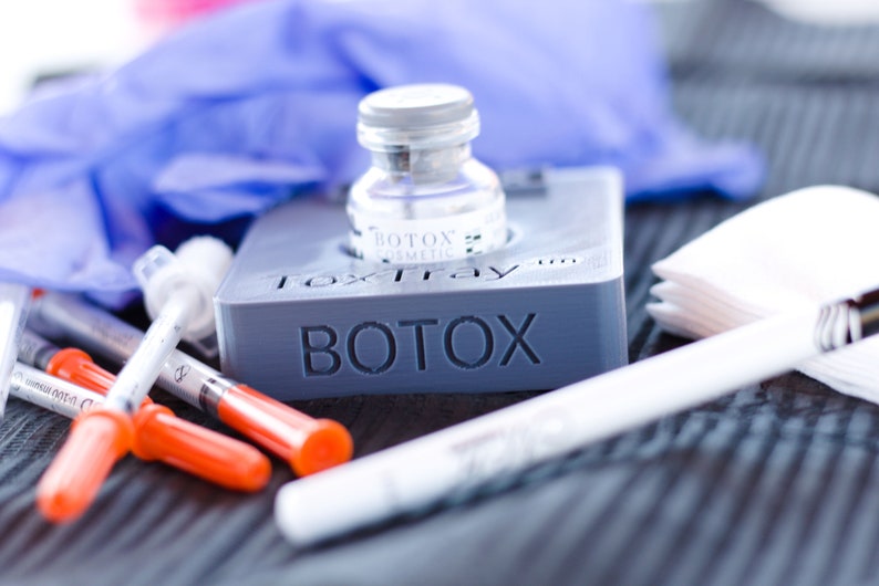 May include: A gray plastic tray with the word "BOTOX" printed on it. A clear glass vial with a blue label that says "BOTOX COSMETIC" is sitting in the tray. There are also several orange and clear syringes, a white cotton swab, and a stack of white paper squares on the tray.