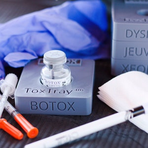 May include: A gray plastic tray labeled "ToxTray™ BOTOX" with a clear glass vial labeled "BOTOX COSMETIC" inside. The tray is next to a stack of white wipes and a blue latex glove. There are also several orange and clear syringes on the table.