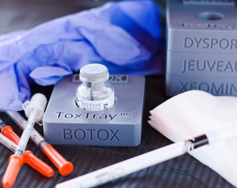 May include: A gray plastic tray labeled "ToxTray™ BOTOX" with a clear glass vial labeled "BOTOX COSMETIC" inside. The tray is next to a stack of white wipes and a blue latex glove. There are also several orange and clear syringes on the table.