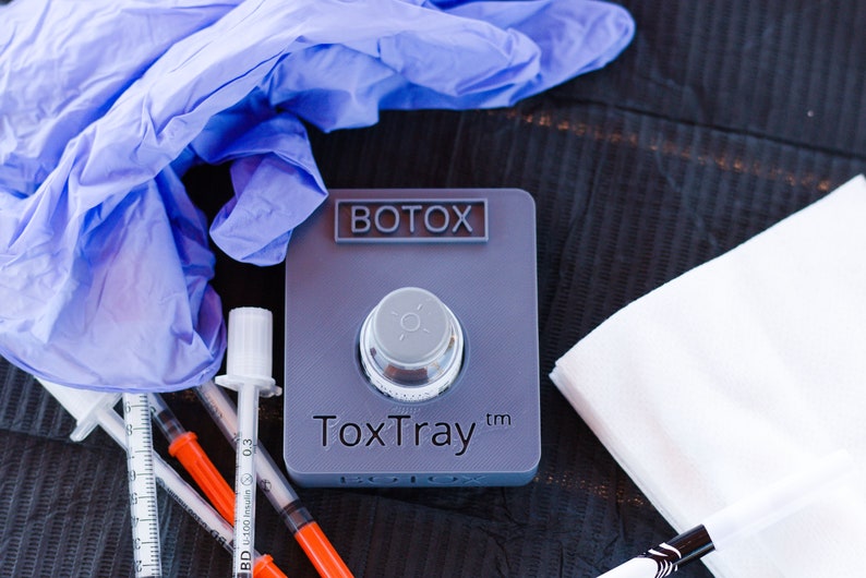 May include: A gray plastic tray with the text "BOTOX" and "ToxTray&trade;" on it. The tray has a small, round, silver container in the center. There are also three syringes, a pair of blue latex gloves, and a white tissue on a black surface.