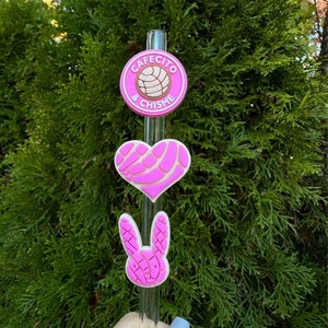 May include: A clear glass straw with three pink and white charms. The top charm is a circle with the text "Cafecito & Chisme" and a bread design. Below is a heart charm, and a bunny charm at the bottom.
