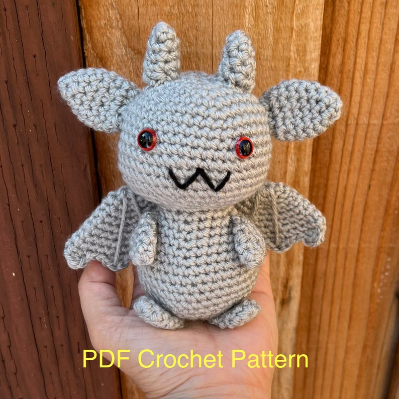 Op de afbeelding: Een gehaakte grijze gargoyle pop met rode ogen, een zwart gestikte mond en kleine vleugels. De pop heeft twee kleine hoorns en grote oren. De tekst "PDF Crochet Pattern" is zichtbaar onderaan de afbeelding.