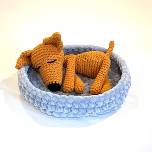 May include: A crocheted dog toy in a light blue crocheted bed. The dog is brown and has its eyes closed. The bed is round and has a soft, fluffy texture.