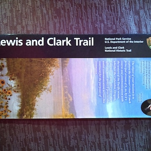 May include: A vintage-style brochure for the Lewis and Clark National Historic Trail. The brochure features a scenic landscape image of a river and trees, with text describing the trail's history and purpose. The text includes the year 1803 and a quote from President Thomas Jefferson.