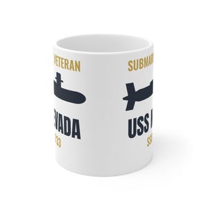 May include: A white ceramic mug featuring a dark blue submarine silhouette, gold text reading VETERAN and SUBMARINE, and dark blue text reading NEVADA 733 and USS NEVADA SSN 733. The design is printed on the exterior surface of the cylindrical mug.