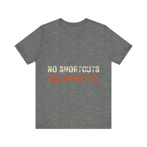 May include: A heather gray short-sleeve t-shirt featuring the text NO SHORTCUTS in a distressed white font and NO REGRETS in a distressed orange font centered on the chest. The garment has a crew neckline and a standard straight hemline.