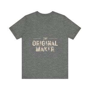 May include: A heather gray short-sleeve t-shirt featuring the text THE ORIGINAL MAKER printed in a distressed, off-white font across the center of the chest. The garment has a crew neckline and a standard fit with a textured fabric appearance.