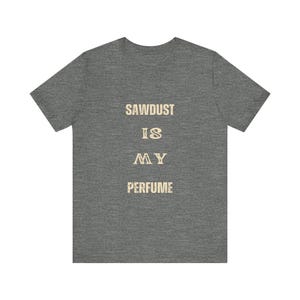 May include: A dark heather gray short-sleeve t-shirt featuring the text "SAWDUST IS MY PERFUME" printed in a distressed, light beige font centered on the chest. The garment has a crew neckline and a standard fit with a textured fabric appearance.