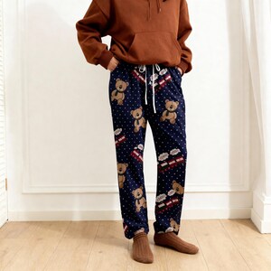 Op de afbeelding: Marineblauwe pyjamabroek met een herhaald patroon van teddyberen met sjaals en kleine rood-witte treinillustraties. De broek heeft een taille met trekkoord en is gemaakt van een zacht materiaal.