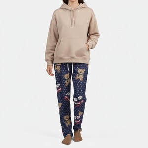 Op de afbeelding: Een beige hoodie met een voorzak en een trekkoord. Het model draagt een marineblauwe pyjamabroek met een patroon van teddyberen, witte stippen en kleine geschenkdozen. De pyjamabroek heeft een trekkoord.