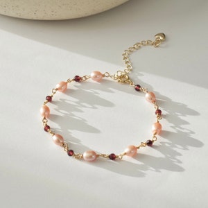 Peut inclure: Un bracelet doré orné de perles roses ovales et de petites perles rondes rouge foncé. Le bracelet est doté d'une chaîne réglable et d'un charm en forme de cœur. Le bijou est présenté sur une surface blanche.