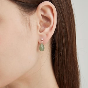 Peut inclure: Gros plan sur une oreille portant une boucle d'oreille en argent avec une pierre précieuse verte pâle en forme de goutte. La boucle d'oreille pend sous le lobe de l'oreille, mettant en valeur la surface lisse et les subtiles variations de couleur de la pierre.