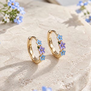 May include: A pair of gold hoop earrings featuring small enamel flower charms in shades of light blue and purple. Each hoop is decorated with three delicate floral accents, showcasing a polished metallic finish and a hinged closure mechanism.