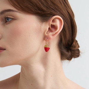 May include: A close-up of a person's ear wearing a gold earring. The earring features a cluster of small, red beads resembling a raspberry, with a gold leaf accent. The earring hangs from a gold hook.