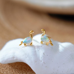 May include: A pair of gold-coloured dinosaur-shaped stud earrings. Each earring features a pearlescent, oval-shaped stone. The earrings are set on a white, textured surface, with a blurred background.