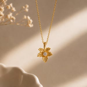 Può includere: Una collana color oro con una catenina sottile e un ciondolo a forma di fiore a cinque petali. Il centro del ciondolo floreale è incastonato con una piccola pietra preziosa trasparente a taglio rotondo che brilla sui petali dorati.