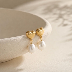 May include: A pair of gold heart-shaped earrings with a white, pear-shaped pearl drop. The earrings are presented on a white surface, with soft shadows in the background. A classic jewellery design.