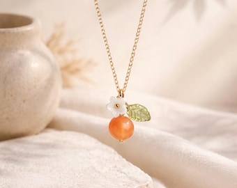 May include: A gold-toned chain necklace featuring a pendant with a round orange bead, a small white five-petaled flower, and a textured green leaf charm. The delicate metal chain supports the cluster of charms, which hang together as a single unit.