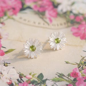 May include: A pair of silver stud earrings shaped like daisies, each featuring a small, faceted, light green gemstone at the centre. The petals are textured with fine lines, radiating outward from the central stone to form a flower shape.