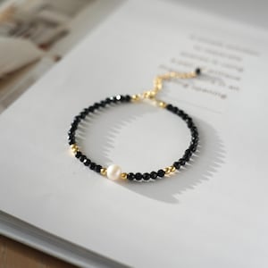 May include: An elegant bracelet with black beads, gold details, and a central white pearl. The bracelet is displayed on a white surface, highlighting its design. This piece of jewellery is a sophisticated accessory.