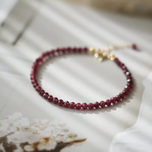 Pu&ograve; includere: Un delicato braccialetto con piccole perline sfaccettate di colore rosso intenso. Le perline sono infilate insieme, formando un cerchio completo. Il braccialetto ha una chiusura e una catena color oro per regolare la taglia. Il gioiello &egrave; esposto su una superficie bianca.
