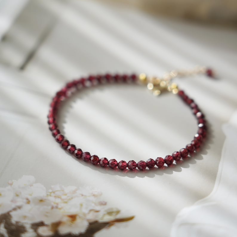 Pu&ograve; includere: Un delicato braccialetto con piccole perline sfaccettate di colore rosso intenso. Le perline sono infilate insieme, formando un cerchio completo. Il braccialetto ha una chiusura e una catena color oro per regolare la taglia. Il gioiello &egrave; esposto su una superficie bianca.
