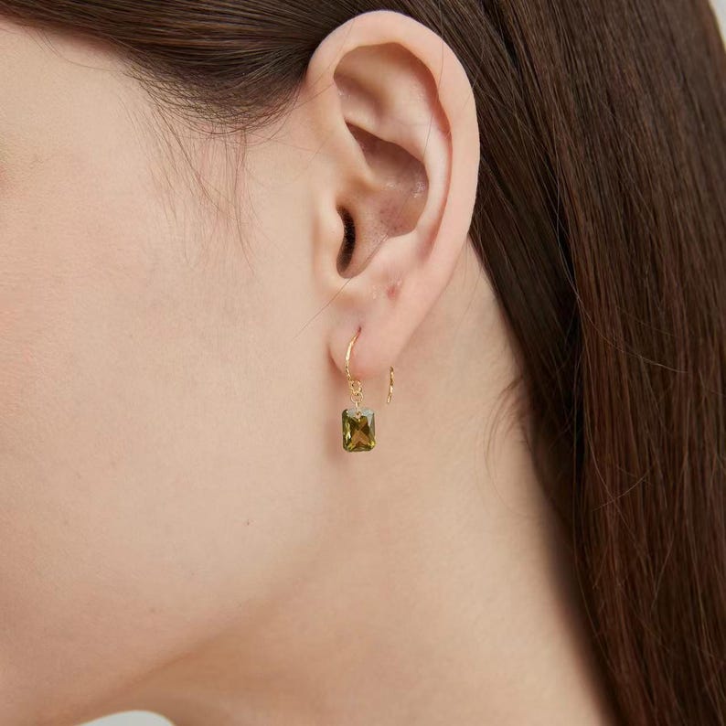 Puede incluir: Primer plano de un pendiente de aro dorado con una gema rectangular verde oliva. El pendiente cuelga de una oreja. El aro dorado es fino y la gema es de un color verde intenso y terroso. El pendiente es una joya sencilla pero elegante.