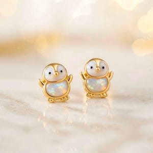 Op de afbeelding: Een paar goudkleurige pinguïn-oorstekers. Elke oorbel heeft een witte opaalbuik, zwarte ogen en uitgestrekte armen. De oorbellen staan tegen een zachte, vage achtergrond.