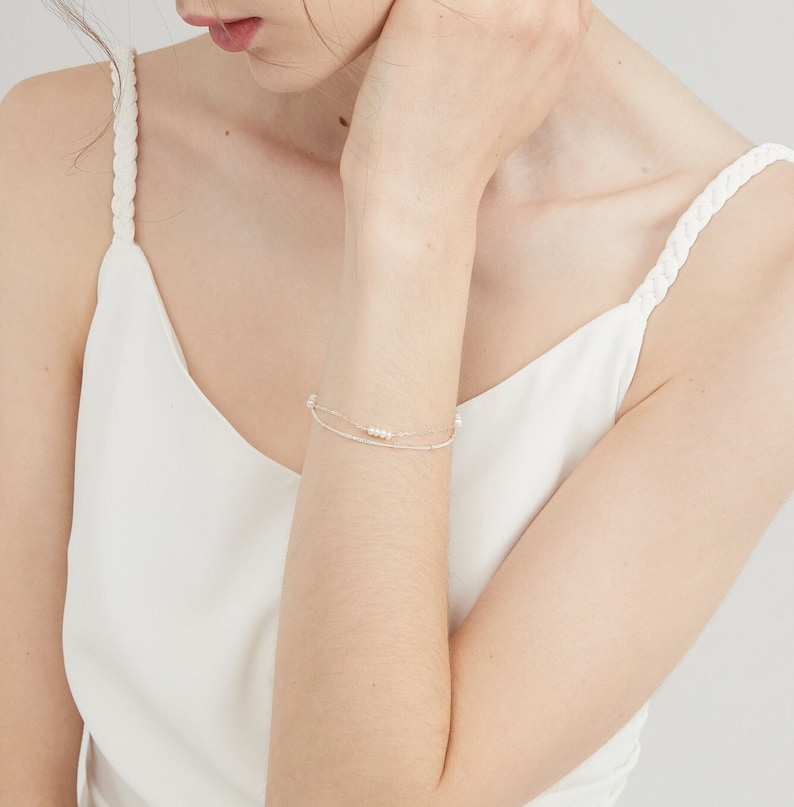 May include: A delicate silver chain bracelet with a row of small white pearls. The bracelet is layered with a thinner silver chain.