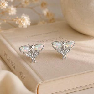 May include: A pair of silver-toned moth-shaped stud earrings featuring iridescent, oval-shaped opal-like cabochons on each wing. The metal wings display intricate engraved line details, and the central body of each moth is slender and metallic.